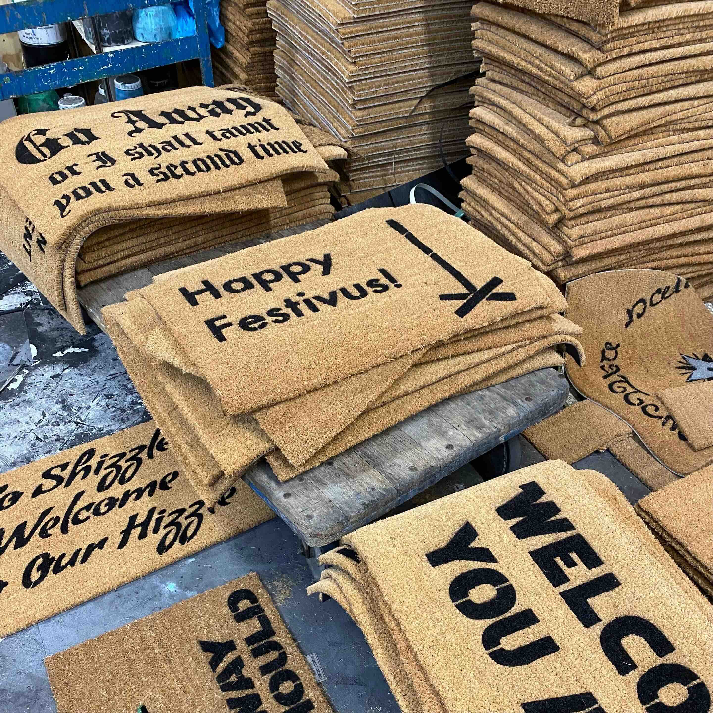 Several funny Christmas doormats laid out together on a workshop floor.
