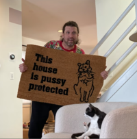 photo of a man showing a doormat reading This house is Pussy protected toa cat