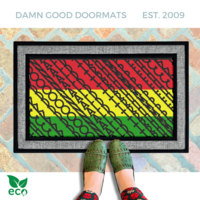 a pair of feet in black and white striped rainboots standing on an all-weather colorful, patterned all weather doormat with a geometric Fuck Donald Trump design in pan-African red, yellow, and green.