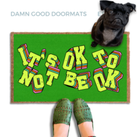 A pair of green shoes and a black pug dog are featured on a green doormat with the text "It's OK to BE OK" written on it.