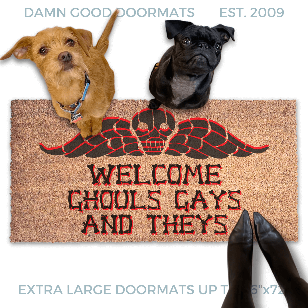 a humorous LGBTQ Halloween door mat with the text "Welcome Ghouls, Gays and Theys" and terrier and a black pug on top of it. There is also a pumpkin and a person's feet visible in the image.
